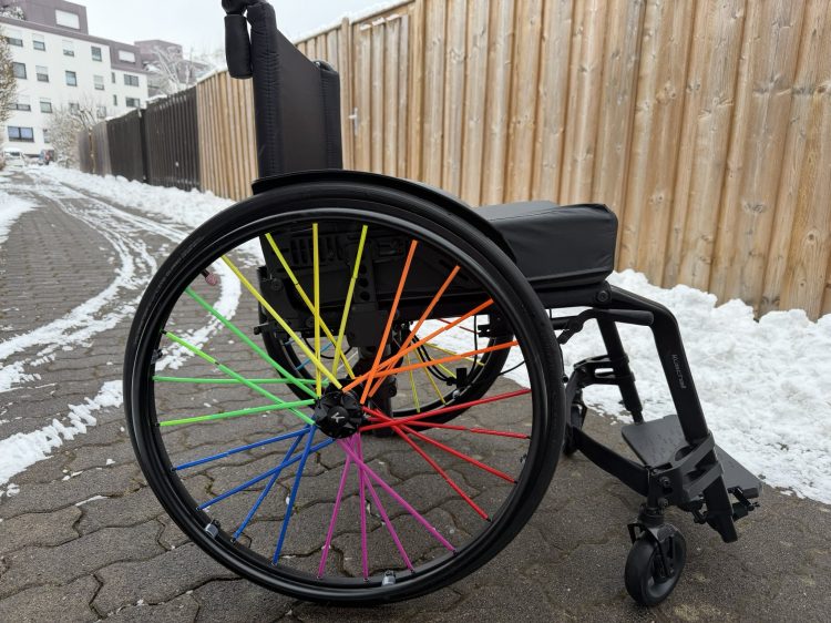 Ein Rollstuhl auf dem Bürgersteig. Er hat bunte Speichen.