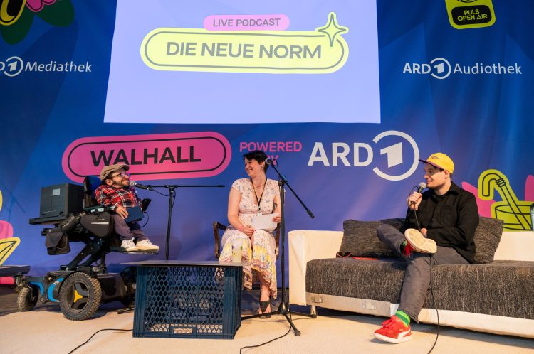Raul sitzt im Elektrorollstuhl, Karina auf einem Stuhl und Jonas auf einem Sofa. Alle sprechen in Mikrofone.