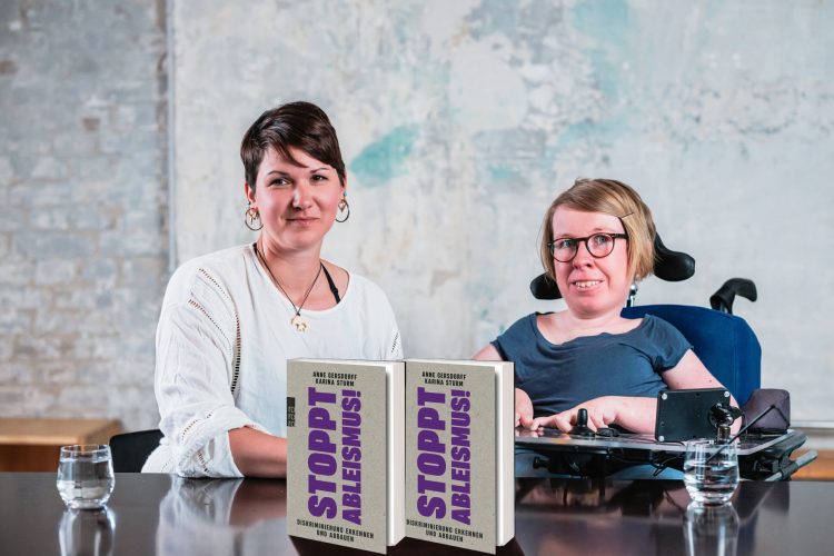 Karina Sturm und Anne Gersdorff sitzen hinter einem Tisch und schauen in die Kamera. Auf dem Tisch aufgestellt sind zwei Exemplare ihres Buches "Stoppt Ableismus".