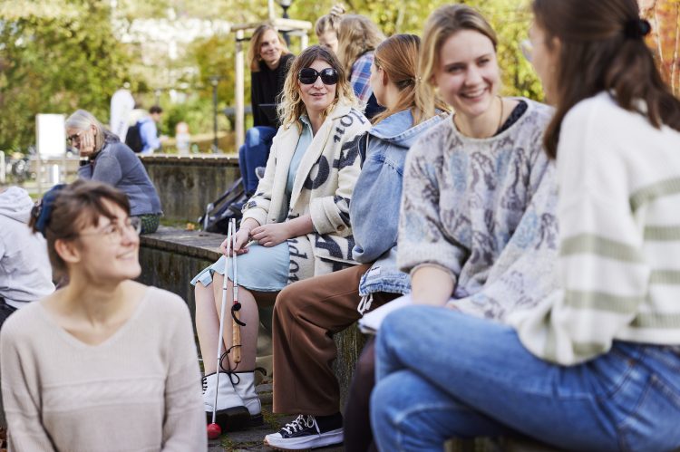 Mehrere Menschen sitzen in einer Gruppe zusammen und unterhalten sich, darunter auch eine Person mit Sonnenbrille und Langstock