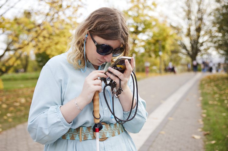 Eine Frau mit Sonnenbrille und Langstock schaut sehr nah auf ihr Smartphone.
