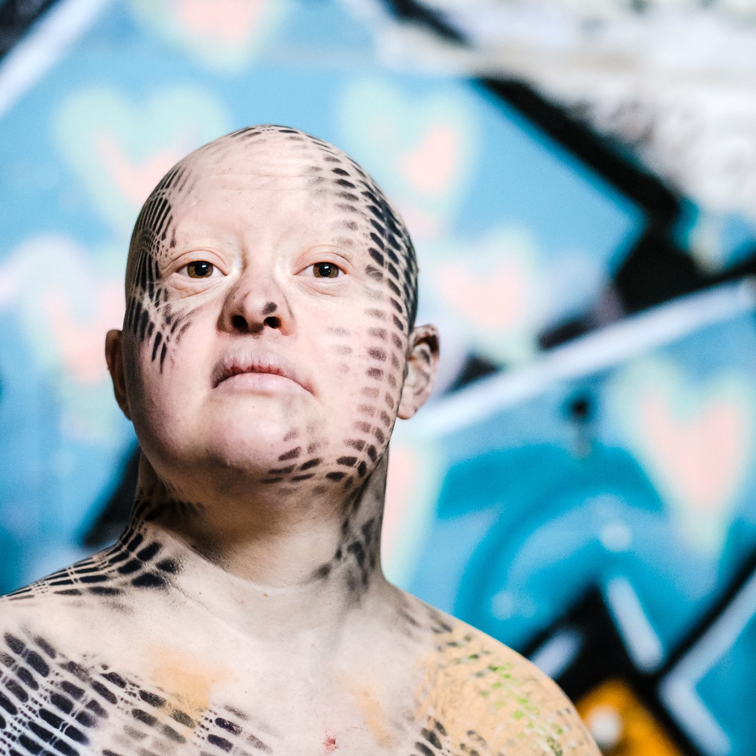 Die Räuber | Theater RambaZamba Zora Schemm, eine Frau mit Down-Syndrom steht vor einem gelb-blauen Graffiti. Sie hat keine Haare und ist schulterfrei bekleideti. Sie ist mit schwarzen Mustern am Körper und Kopf bemalt.